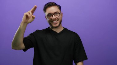 Sakallı, gözlüklü, parmakları mor arka planda selam veren, günlük siyah gömlek giyen, stüdyo ortamında dostane ve cana yakın bir jest yapan genç bir adam..