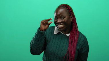 Pembe örgülü bir kadın yeşil süveterle yeşil arka plana karşı küçük bir jest yapıyor, gülümsüyor ve kameraya bakıyor, mizah ve pozitiflik taşıyor..