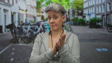 Kısa saçlı, gri kapüşonlu ve kaldırım taşı kaldırımlı bir sokakta kollarını çapraz bağlayan bisikletli bir kadın..