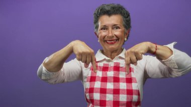 Gri saçlı, kırmızı kareli önlük giyen son sınıf öğrencisi bir kadın canlı mor bir arkaplan karşısında neşeli ve çekici bir ifade sergiliyor..