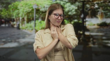 Young blonde woman in beige shirt stands in green outdoor park holding her neck with both hands; discomfort.