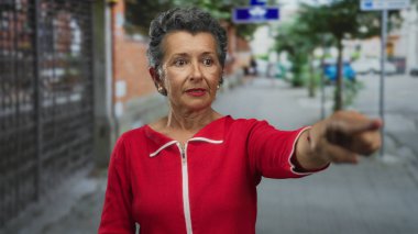 Son sınıf öğrencisi, gri saçlı, kırmızı elbiseli bir kadın gün boyunca şehir caddesini işaret ediyor..