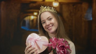 Taç giyen kadın kalp şeklinde hediye kutusu ve pembe çiçekleri bina koridorunda tutuyor. Aşk romantizmi..