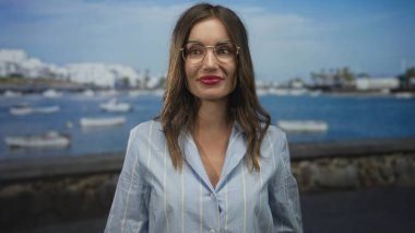 Gözlüklü kadın, açık mavi gömlek giyen güneşli bir sahil limanında demirli beyaz tekneler tarafından kameraya geniş bir şekilde gülümsüyor; mutluluk..