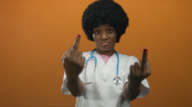 Beyaz önlük ve steteskop giyen genç Afro-Amerikan kadın doktor, turuncu stüdyoda orta parmağını kaldırıyor..