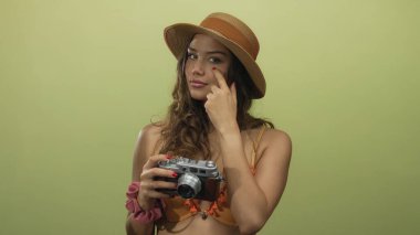 Kameralı bikinili kadın sarı arka planda poz veriyor, şapka takıyor ve turist gibi davranıyor, yaz macerasına dair ipuçları sunuyor..