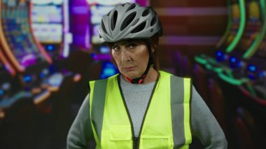 Kumarhane ortamında kask takan yaşlı bir kadın kumar makineleri görünür ve parlak ışıkların altında varlığını vurgulayan bir güvenlik yeleği canlı bir atmosferi işaret ediyor..