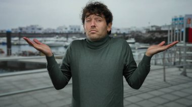 Limanda duran bir adam, süveter içinde yakışıklı görünüyor, arka planda botlar, marinaya karşı düşünceli bir ifadeyle sakin bir sahne yaratıyor..