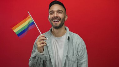 Genç İspanyol adam gülümsüyor ve renkli bir lgtb bayrağını canlı kırmızı bir arkaplana tutuyordu. İfade dolu bir portrede gurur ve neşe saçıyordu..