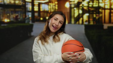 Sokakta elinde basketbol topuyla gülümseyen genç bir kadın aydınlanmış arka planda, açık hava kentsel ortamının tadını çıkarıyor..