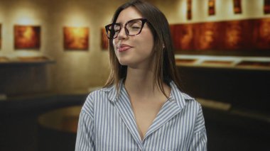 Gözlüklü ve çizgili gömlek giyen kadın, soyut tabloyu izlerken müze galerisinde gözlerini kapatıyor; sükunet yansıması hayranlığı.