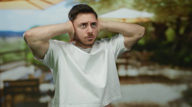 Beyaz tişörtlü, sakallı genç bir adam iki eliyle güneşli bir terasta açık hava restoranında stres ya da hüsranı öneriyor.