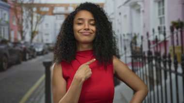 Kırmızı bluzlu ve kıvırcık saçlı genç bir kadın demir çitlerle ve park edilmiş arabalarla dolu bir sokakta kendini işaret ediyor..