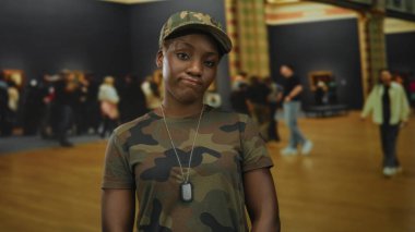 Kamuflaj üniformalı ve şapkalı kadın künye takıyor dudaklarını bastırıyor ve müze binasına bakıyor..