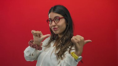 Beyaz bluz ve kırmızı gözlük takan genç İspanyol kadın parmaklarını şakacı duruşu olan kırmızı bir stüdyo duvarına doğru gösteriyor..
