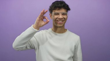 Gözlüklü ve beyaz kazaklı bir adam mor bir stüdyoda işaret dilini kullanırken geniş bir gülümseme sergiliyor..