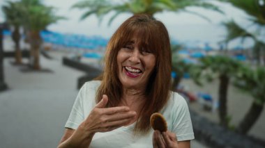 İspanyol kıdemli kadın sahilde kurabiye yiyor. Plaj arka planında palmiye ağaçları var..