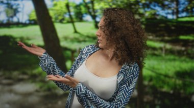 Orman parkında çıplak göğüs ve avuçlarını gösteren bir kadın sorgulama duruşuyla yana bakıyor; belirsizlik..