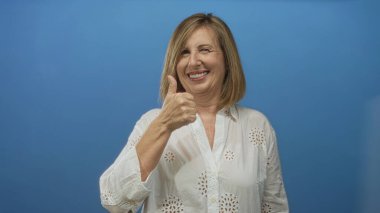Orta yaşlı bir kadın, stüdyoda mavi duvara karşı beyaz bluz giyen, iyimser, kendine güveni yüksek bir jest gösteriyor..