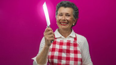 Kareli önlüklü yaşlı kadın elinde bıçakla canlı pembe arka plana gülümsüyor, neşeli yemek pişirme deneyimi yayıyor..