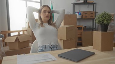 Yeni evinde dizüstü bilgisayarla çalışan bir kadın. Etrafı parlak oturma odasındaki kutuları taşımakla çevrili. Yerleşmeye ve yerleşmeye odaklanmış.
