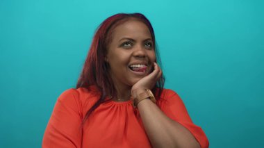 Turkuaz bir stüdyo ortamında el ele gülümseyen ve turuncu bluz giyen bir kadın..