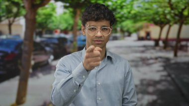 Mavi gömlekli ve gözlüklü adam şehir kaldırım ortamının ortasında kamerayı işaret ediyor; özgüven odaklı.