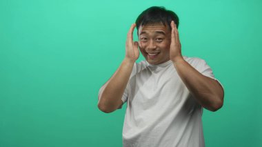 Genç Çinli adam yeşil stüdyoda elleri ile çerçeve gülümsüyor, doğrudan objektife bakıyor; merak hayal gücü yaratıcılığını hayrete düşürüyor.