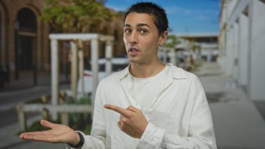 Beyaz gömlek giyen genç İspanyol adam elini uzatır ve güneşli bir sokakta onu işaret eder; güpegündüz rehberlik merakını davet eder.