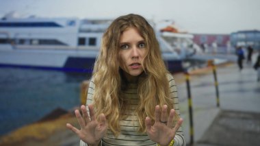 Sıradan sahnelerde kalabalık bir limanda elini kaldıran bir kadın görülüyor. Sarı saçları, feribot iskelesinde çizgili bir süveteri çerçeveliyor..