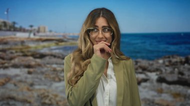 Mavi deniz kıyısındaki güneşli kayalık sahil şeridinde ağzı kaplayan iş ceketli ve gözlüklü bir kadın. Sürpriz merak..