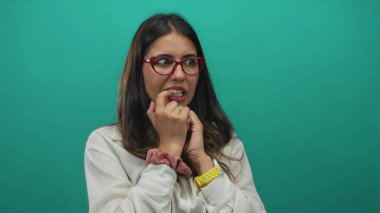 Kırmızı gözlüklü, beyaz bluzlu ve sarı saatli bir kadın stüdyoda tırnak yiyor ve yeşil duvarlı, endişeli bir ifade takınıyor..