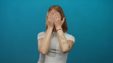 Sarışın genç kadın, mavi arka plan duvarına karşı elleri ile şakacı bir şekilde yüzünü gizliyor..