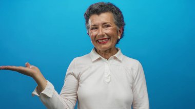Gri saçlı ve pembe bluzlu gülümseyen yaşlı bir kadın canlı mavi bir arka plana karşı açık elle poz veriyor. Stüdyoya mutluluk ve güven saçıyor..