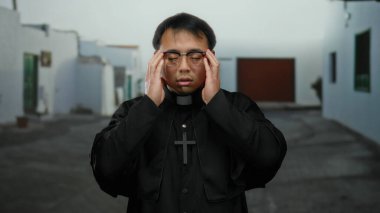 Gözlüklü ve haçlı siyah cüppeli Asyalı bir adam şehir caddesinde duruyor, sakin bir kasaba ortamında düşünce ve yansıma hissi yayıyor..