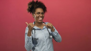 Açık mavi gömlekli, gözlüklü, steteskop takan genç bir doktor pembe stüdyoda gülümseyip başparmak kaldırıyor..