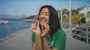 Kadın deniz kenarındaki gezinti güvertesinde yeşil tişört giyip arka planda dağlar olan jestler yaparak açık gökyüzü altında neşeli duygular ifade ediyor..