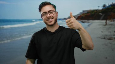 Kumsalda sakallı genç bir adam, açık gökyüzünün altında deniz kenarında el kol hareketi yapıyor. Açık hava ortamında karışık duyguları yansıtıyor..