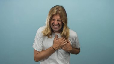 Açık mavi stüdyoda uzun saçlı, terleyen, yüzünü buruşturan ve göğsünü sıkan sarışın bir adam..