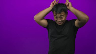 Gülümseyen ve tavşan kulağı yapan bir adam mor bir stüdyoda elleri ile siyah bir tişört giyerek hareket eder..