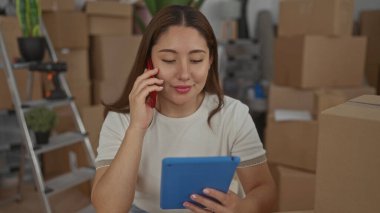 Binada akıllı telefonla konuşurken elinde tablet tutan genç İspanyol kadın, odaklanmış planlama.