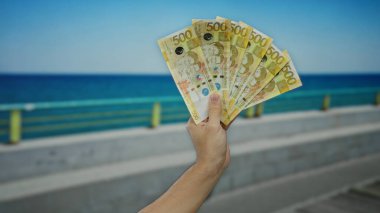 Arka planda açık mavi gökyüzü olan deniz kenarındaki gezinti alanının yanında Filipinli pesoları tutan adam plajda rahatlamış bir havayla para sergiliyor..