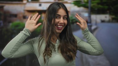 Açık havada gülümseyen, elleri heyecan içinde bulanık bir arkaplana doğru yükselen, neşeli, genç, İspanyol bir kadının doğal ortamında sevinç ifadesi gösteren bir kadın..