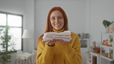 Kızıl saçlı kadın katlanmış giysiler tutuyor. Parlak bir çamaşır odasında gülümsüyor. Sıcak bir ev atmosferi var..