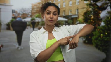 Kadın, süslenmiş noel ağaçları ve bulanık yayaların yakınlarındaki kalabalık bir sokakta, limon rengi üst ve beyaz gömlek giymiş, parmak bileğe işaret ediyor..