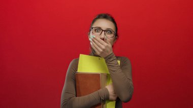 Gözlüklü bir kadın, kalın kırmızı bir duvarın önünde renkli klasörler tutarken ağzını şaşkınlıkla kapatıyor. Duygularını ve gizemini canlı bir şekilde aktarıyor..