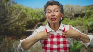 Gri saçlı ve önlüklü yaşlı kadın yeşil ağaçlar ve bitkilerle dolu canlı bir parkta duygusal hareketler yapıyor..