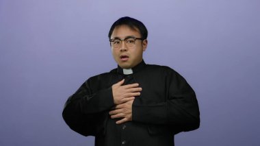 Papaz kıyafetli genç adam, izole bir mor arka planda, kaldırılmış eller ve kaldırılmış gözlüklerle, şok ve dini aktararak irkildi..