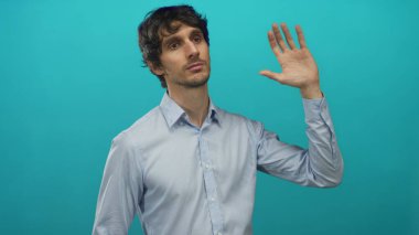 Açık mavi bir gömlek giymiş, mavi bir stüdyo duvarına karşı çıplak elini kaldıran ve portre çekimi bir avuç hareketi gösteren bir adam;.