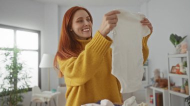 Evde, parlak bir çamaşırhanede elbiselerini katlayan bir kadın. Sarı bir kazak giyiyor. Gülümsüyor ve tatmin olduğunu gösteriyor..